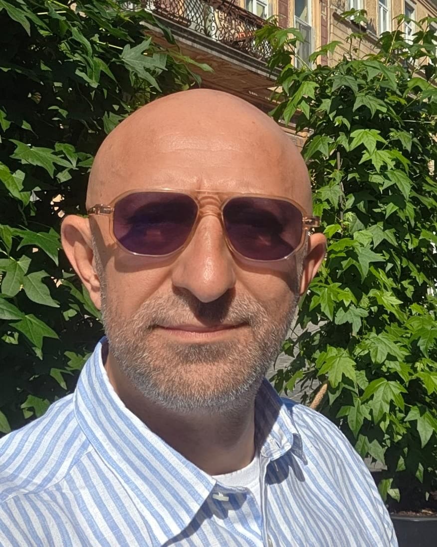 Alexandru Dumitrescu, cu ochelari de soare și cămașă în dungi albastre, în fața unui perete de iederă.