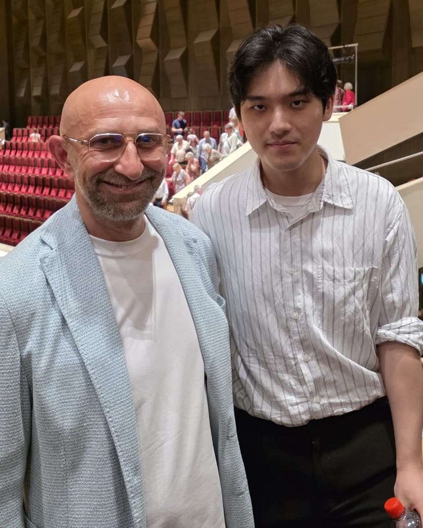 Alexandru Dumitrescu, zâmbind, alături de pianistul Yunchan Lim, în sala Gewandhaus, cu scaunele roșii și peretele de lemn în fundal.